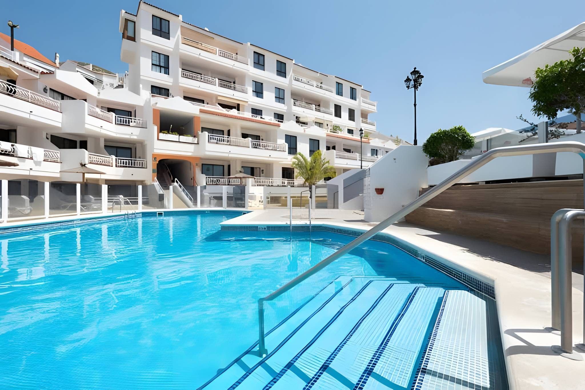 Une piscine à l’eau d’un bleu cristallin, entourée d’un bâtiment blanc doté de balcons et d’un escalier menant à un niveau supérieur.