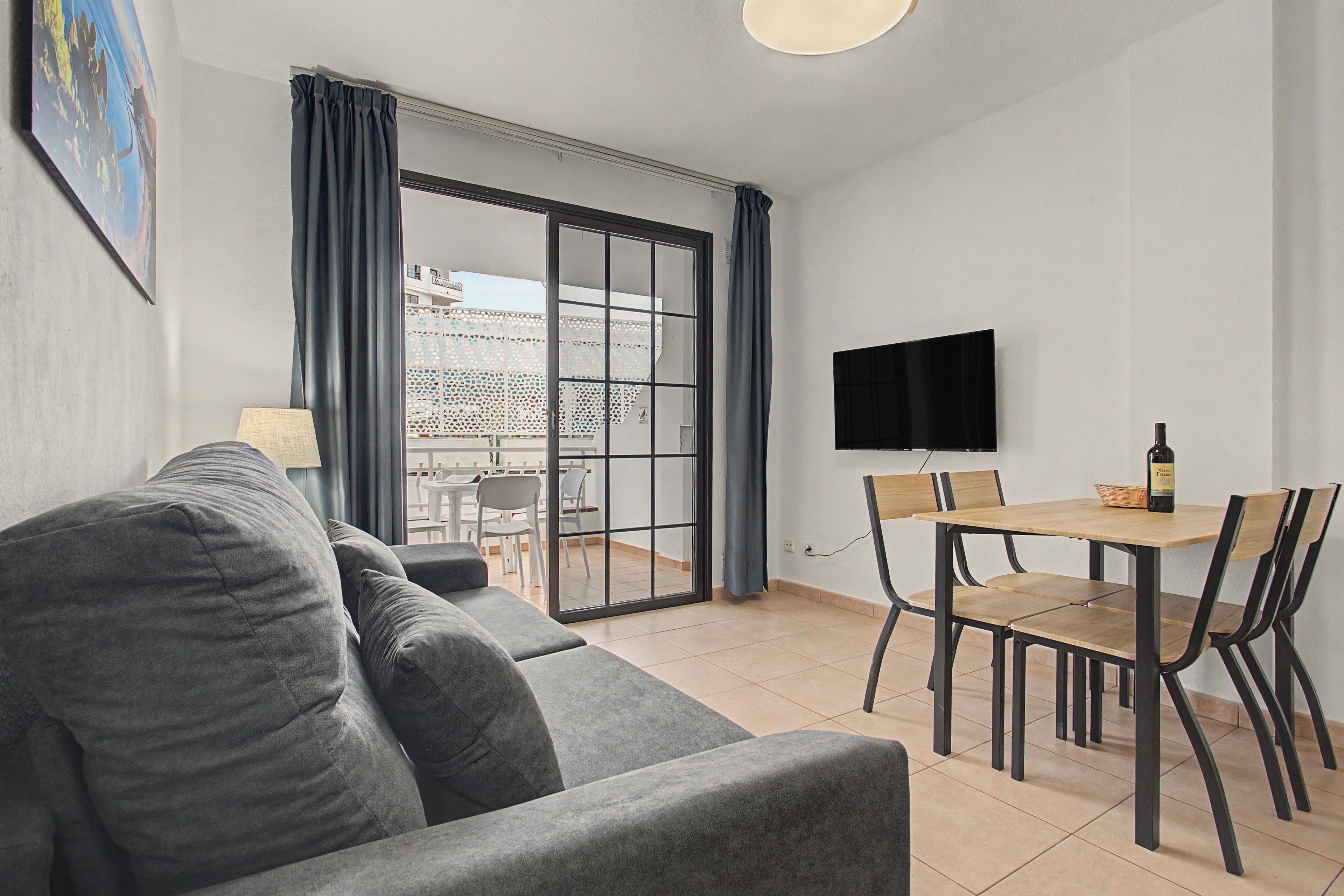 A modern living room with a grey sofa, a dining table with chairs, a flat-screen TV mounted on the wall, and a sliding glass door leading to an outdoor area.