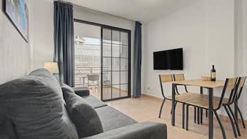 A modern living room with a grey sofa, a dining table with chairs, a flat-screen TV mounted on the wall, and a sliding glass door leading to an outdoor area.