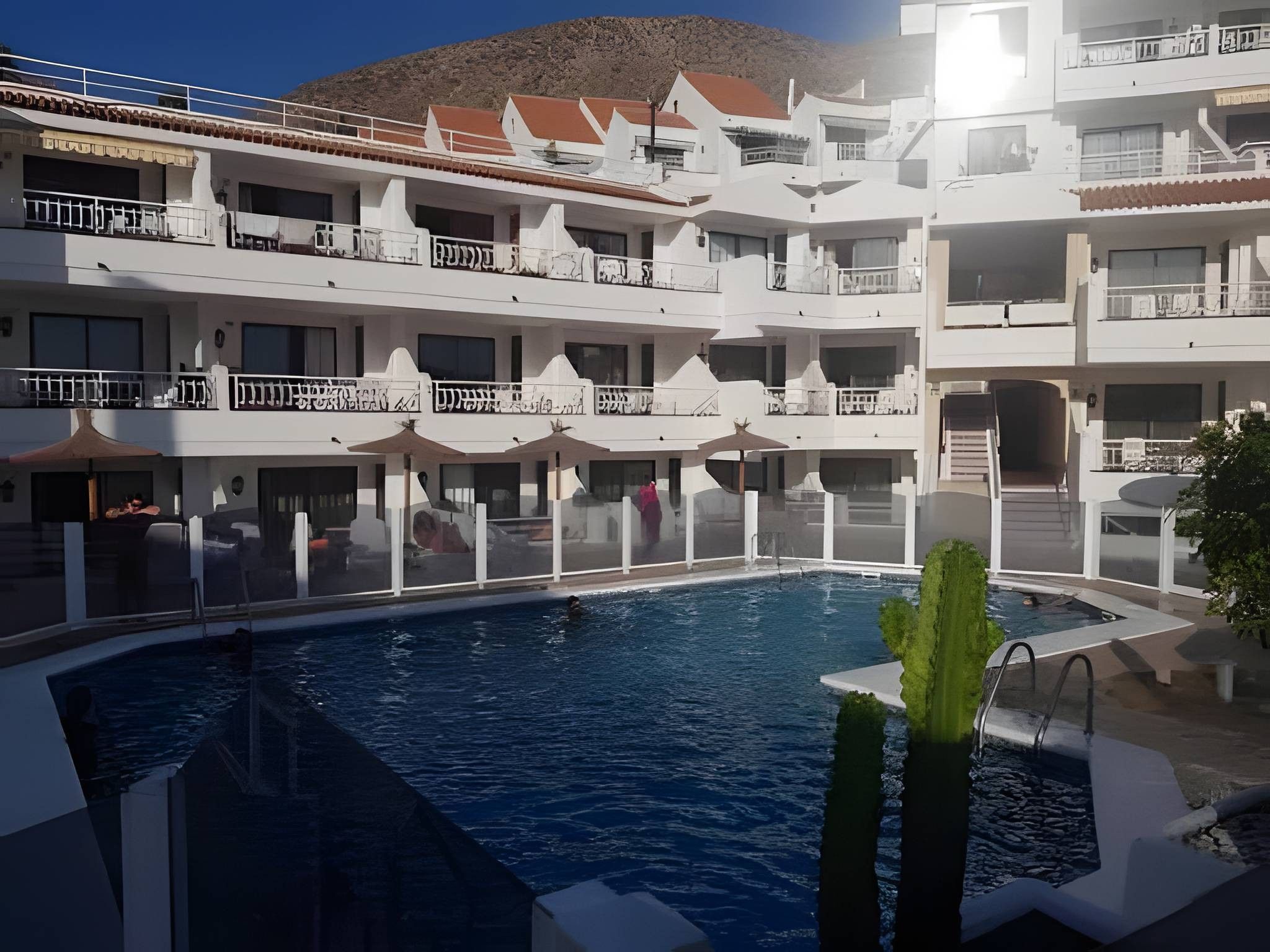 A hotel with a swimming pool, multiple balconies, and a mountainous backdrop.