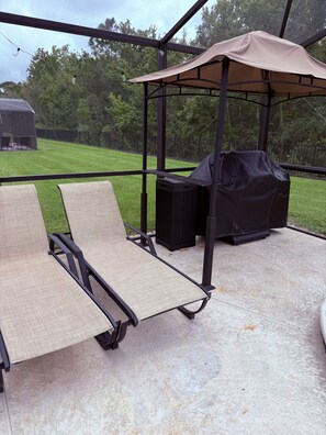 A covered outdoor seating area with two lounge chairs, a barbecue grill, and a trash bin.