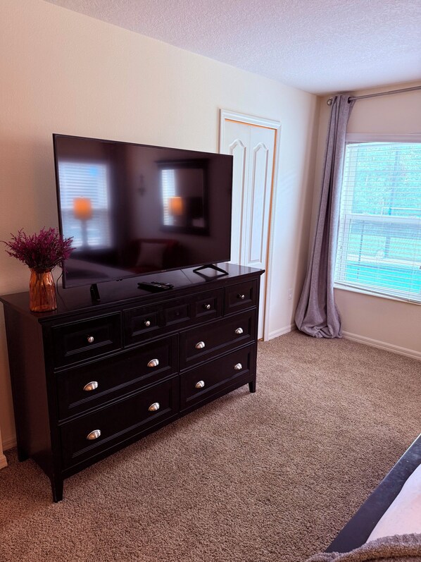 Ein Hotelzimmer mit einem großen Flachbildfernseher auf einer schwarzen Kommode, einer Vase mit lila Blumen und einem Fenster mit Blick auf Grünflächen.