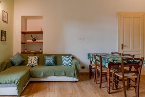 A cozy living room with a green sofa, wooden chairs, a dining table with a green tablecloth, and a white door. - In the vibrant heart of Turin, this flat is all about atmosphere: a spacious, light-filled living room with a fully equipped kitchen, two double bedrooms, a sofa bed and two balconies (one overlooking Largo Montebello, the other a more intimate internal  (Torino)