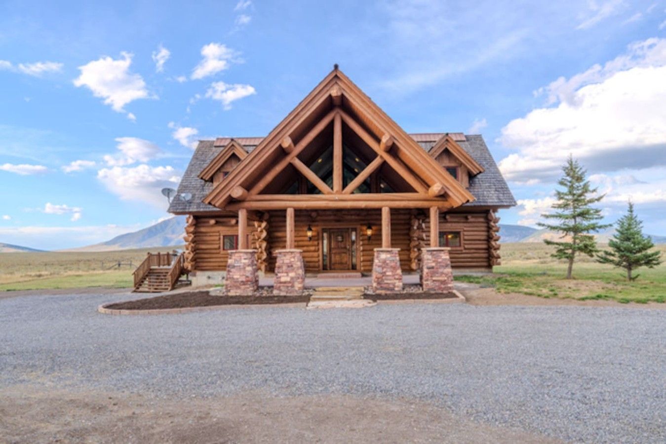 A log cabin with a prominent front porch, a staircase leading to the entrance, and a gravel driveway.