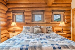 Un dormitorio tipo cabaña de madera con una cama, dos lámparas de noche y tres ventanas.