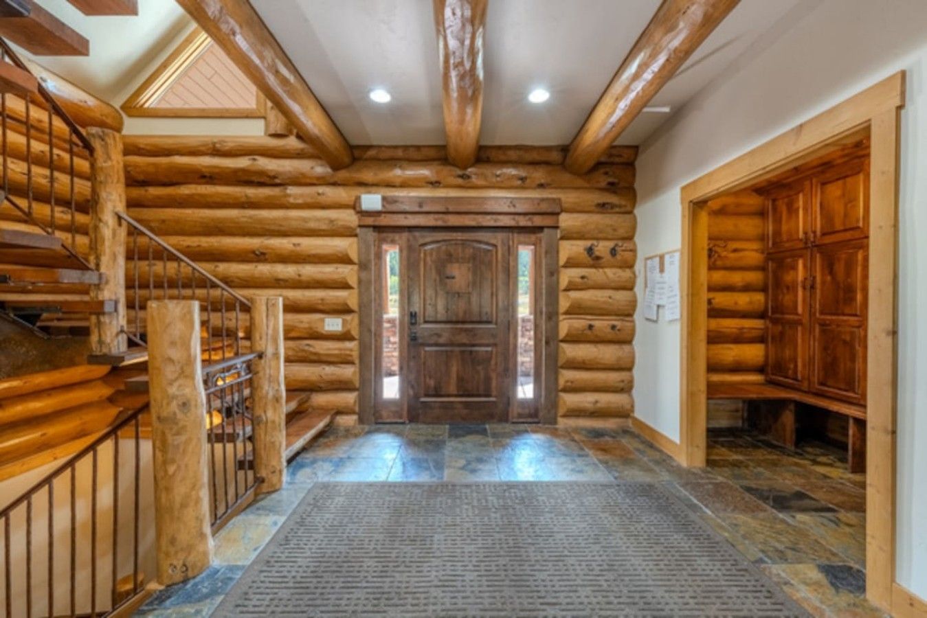 A log cabin interior with a wooden staircase, a central door, and a stone-tiled floor.