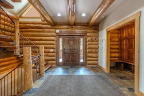 A log cabin interior with a wooden staircase, a central door, and a stone-tiled floor. - Yellowstone Pointe Lodge - Sleeps 30, 20minToYellowstone, Movie Room, Games! (Island Park)