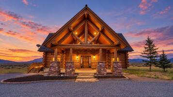 A log cabin with a prominent front porch, a stone foundation, and a mountainous backdrop.