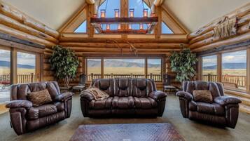 A log cabin interior with a high ceiling, leather sofas, and large windows offering a view of the outdoors.