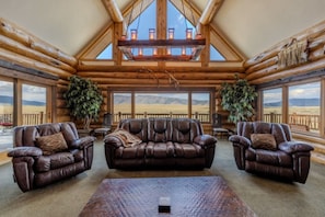 A log cabin interior with a high ceiling, leather sofas, and large windows offering a view of the outdoors. - Yellowstone Pointe Lodge - Sleeps 30, 20minToYellowstone, Movie Room, Games! (Island Park)