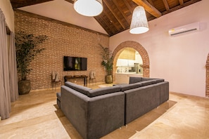 A modern living room with a sectional sofa, a flat-screen TV mounted on a brick wall, and a hanging light fixture. - 11 Bedroom Villa in the Old City (Cartagena de Indias)