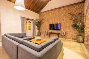 A living room with a sectional sofa, a wooden coffee table, a flat-screen TV mounted on a brick wall, and two potted plants. - 11 Bedroom Villa in the Old City (Cartagena de Indias)