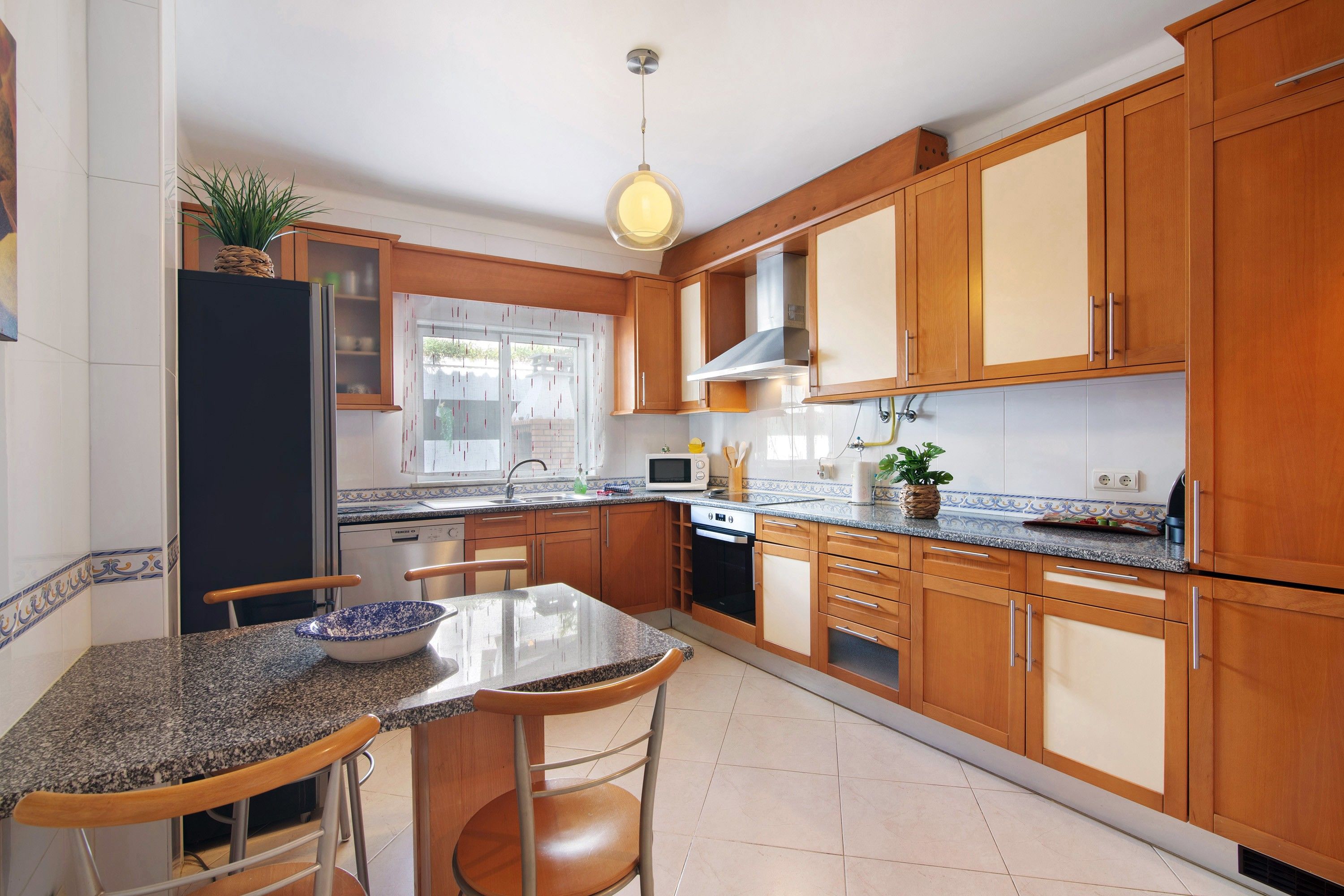 A modern kitchen with wooden cabinets, a granite countertop, a dining area with a table and chairs, and a hanging light fixture.