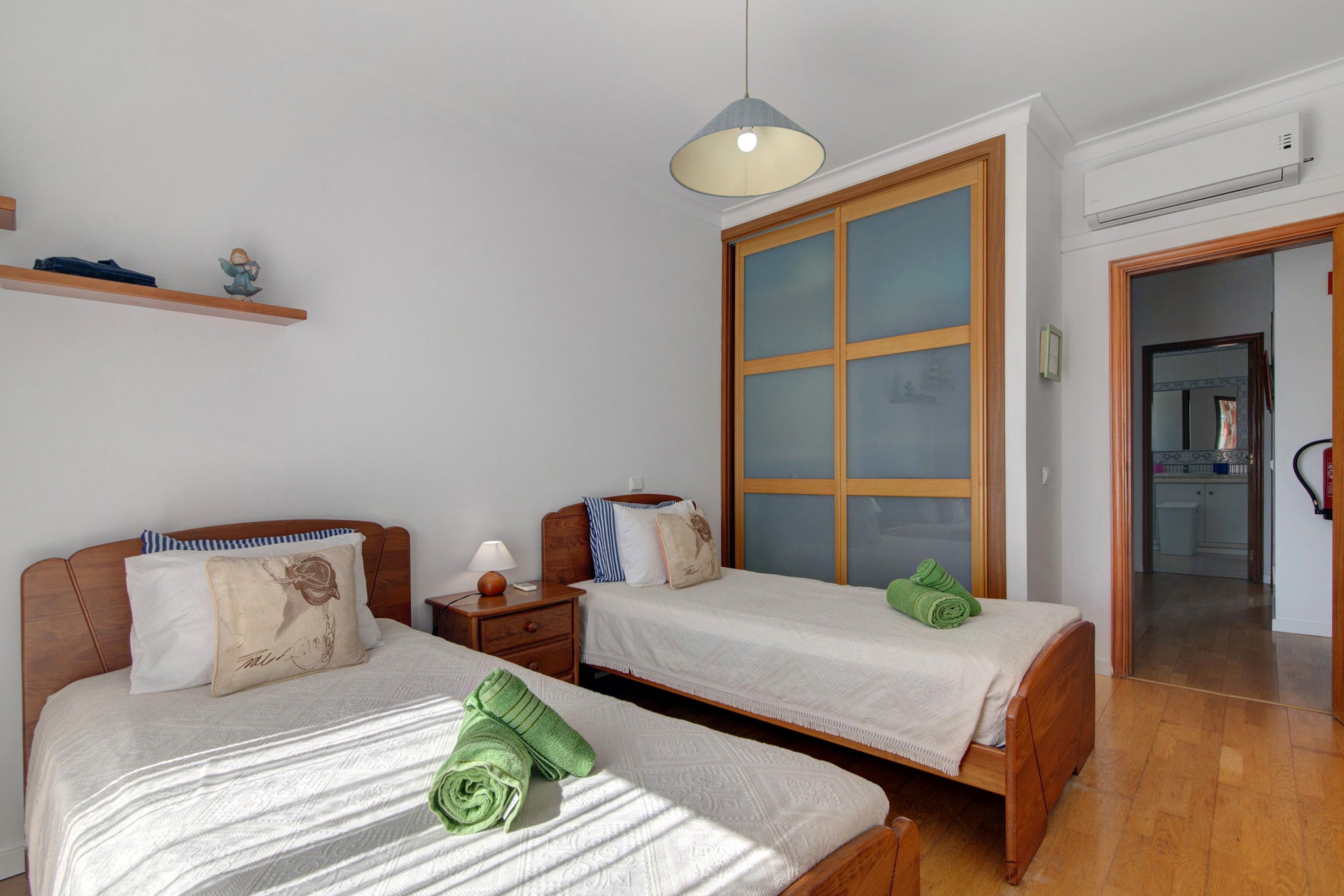 Een kamer met twee bedden, een nachtkastje, een hanglamp en een groot raam met zonwering.