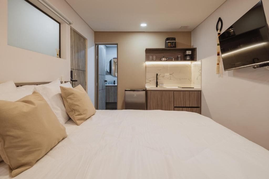 Habitació d'hotel amb llit, petita cuina, mirall i televisió.