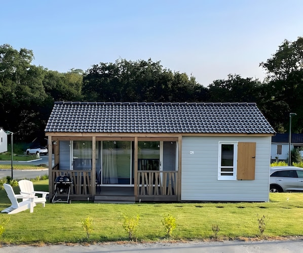 Une petite maison à un étage, avec un toit en tuiles, une véranda avec des chaises et une cour gazonnée.