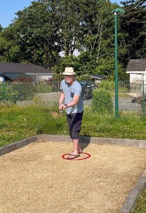 A person playing shuffleboard on a wooden floor with a chain-link fence and greenery in the background.