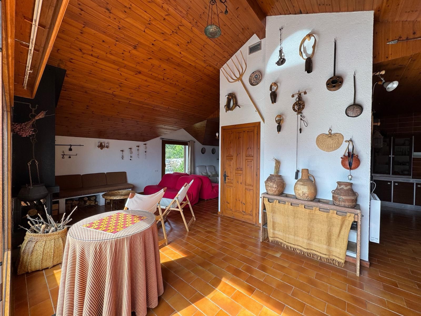 Um interior rústico com teto de madeira, área de jantar com mesa e cadeiras, um sofá e uma parede decorada com utensílios de cozinha e cerâmica.