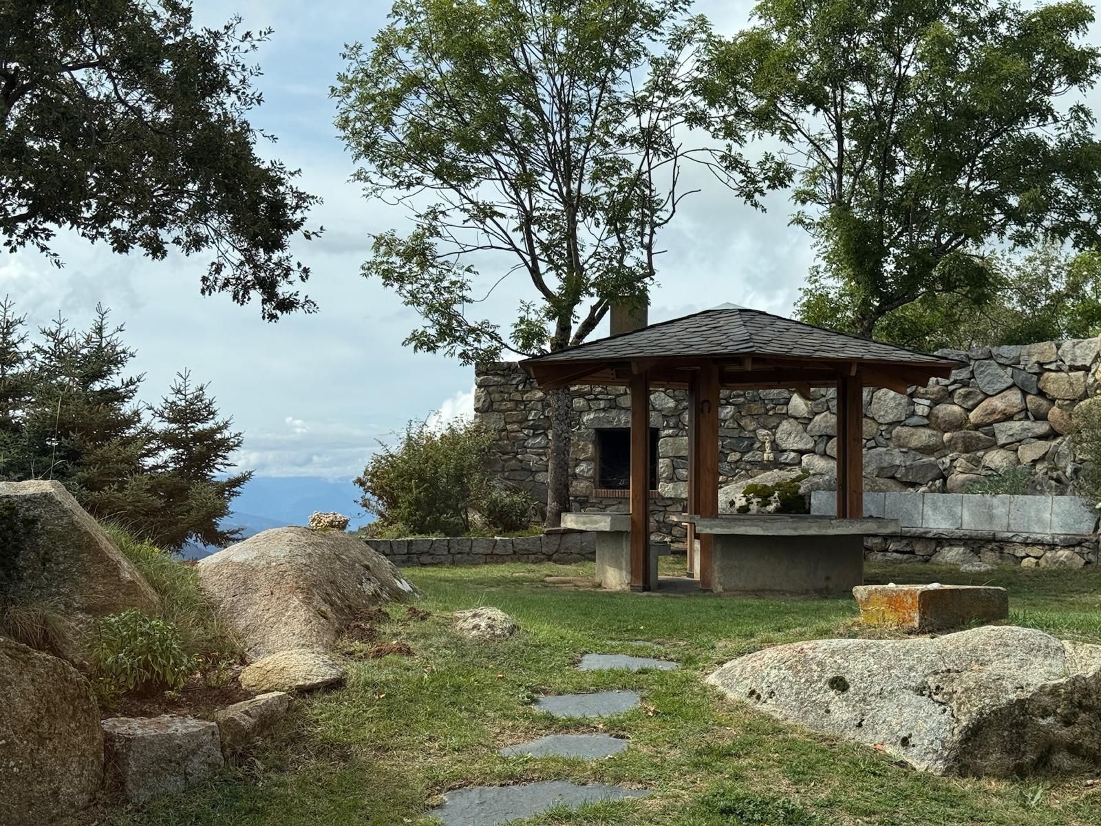 Um gazebo de pedra com telhado de madeira, rodeado por pedras grandes e grama, com árvores e um muro de pedra ao fundo.