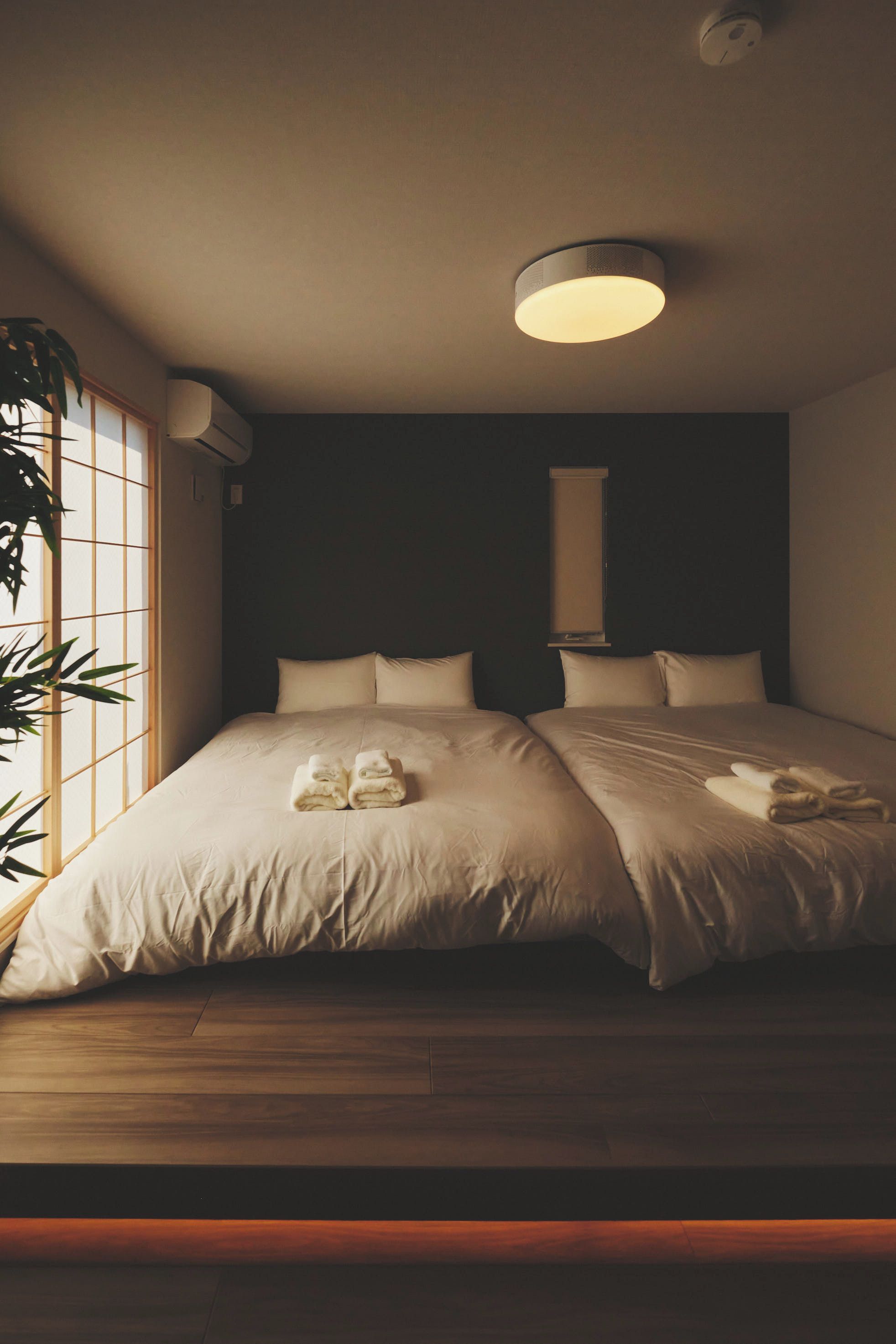 A bedroom with two beds, a window with blinds, and a ceiling light fixture.