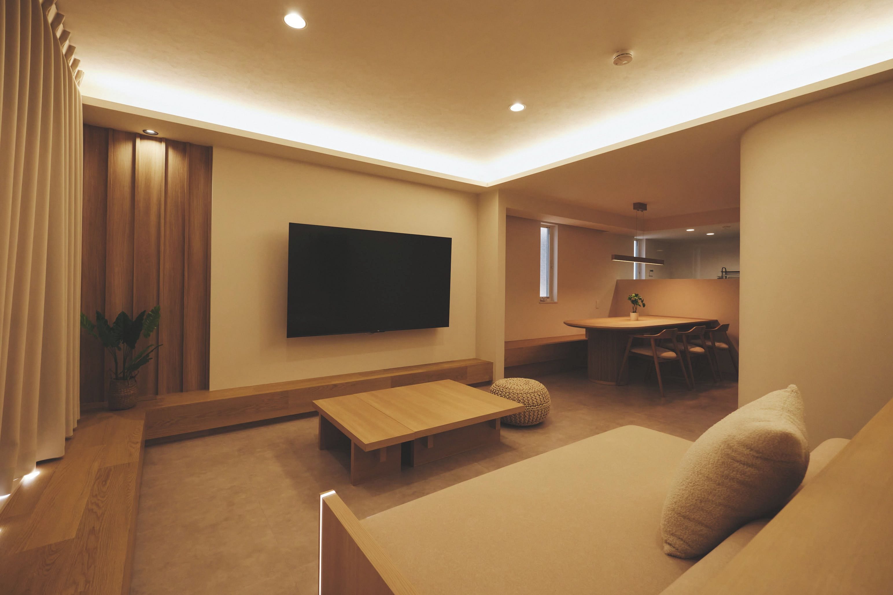 A modern living room with a flat-screen TV, a sofa, a coffee table, and a dining area with a table and chairs.