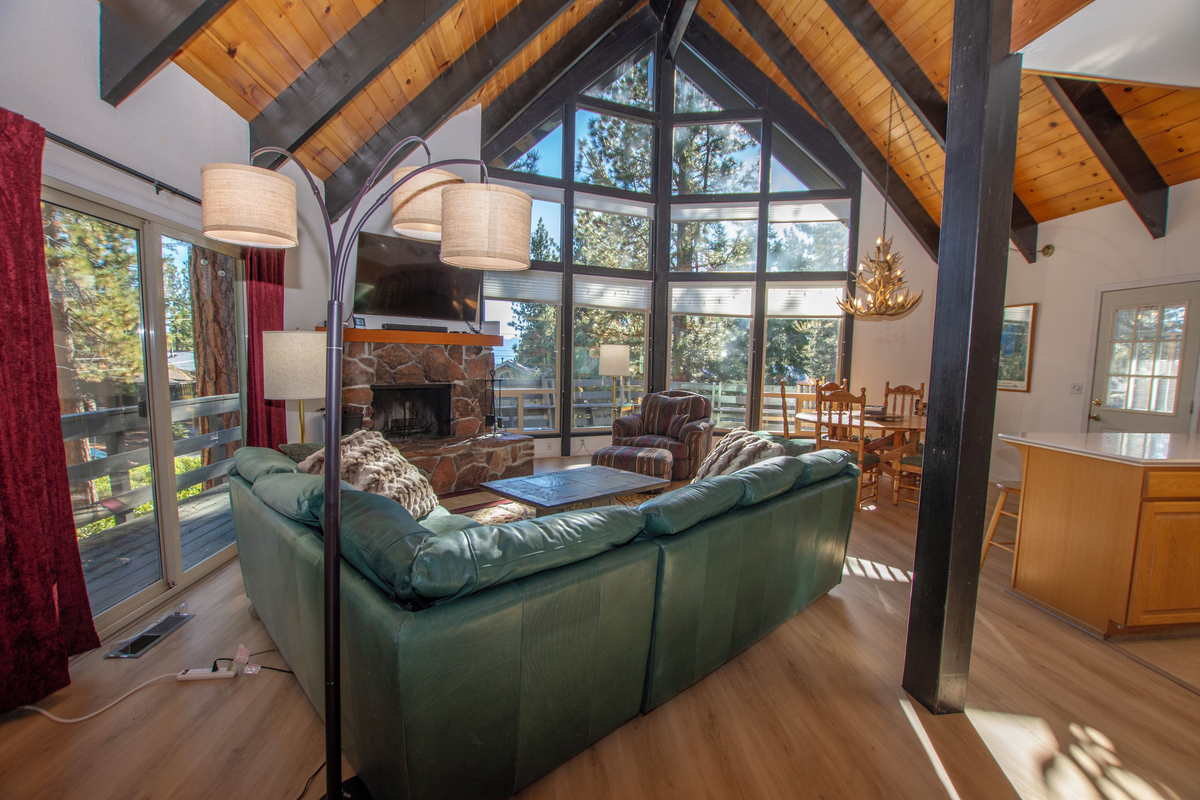 A spacious living room with a stone fireplace, large windows, and a high ceiling with exposed beams.
