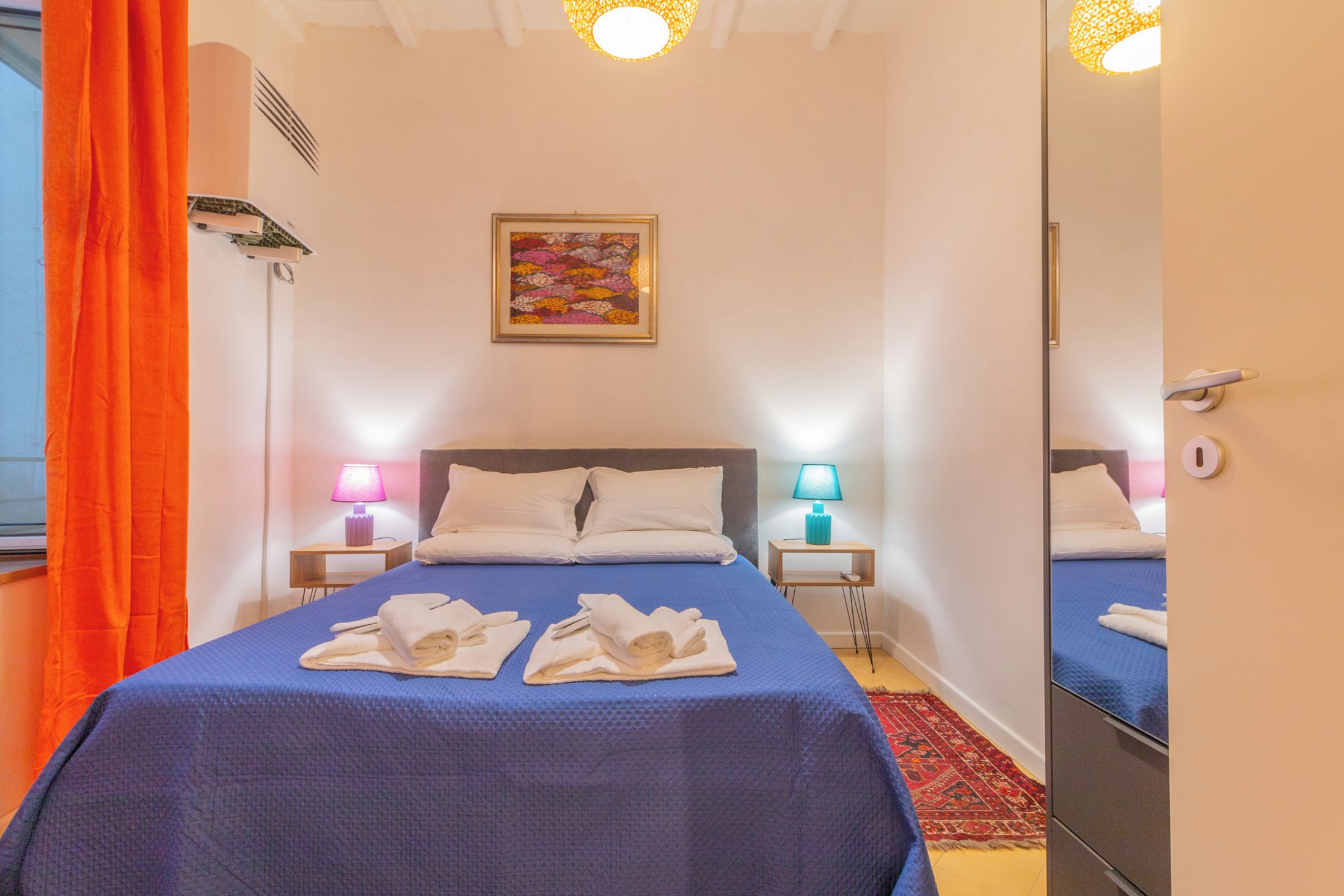 A hotel room with a bed, two bedside lamps, a painting, and a mirror.