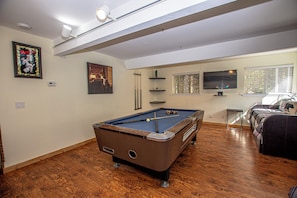 Ein Billardtisch in einem Raum mit Holzboden, einem an der Wand befestigten Fernseher und einem Bett mit Karoüberwurf.