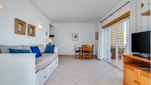 A living room with a sofa, a dining table with chairs, a TV, and a window with a view. - 2BR Flat with Balcony & Shared Pool by LovelyStay (Portimão)