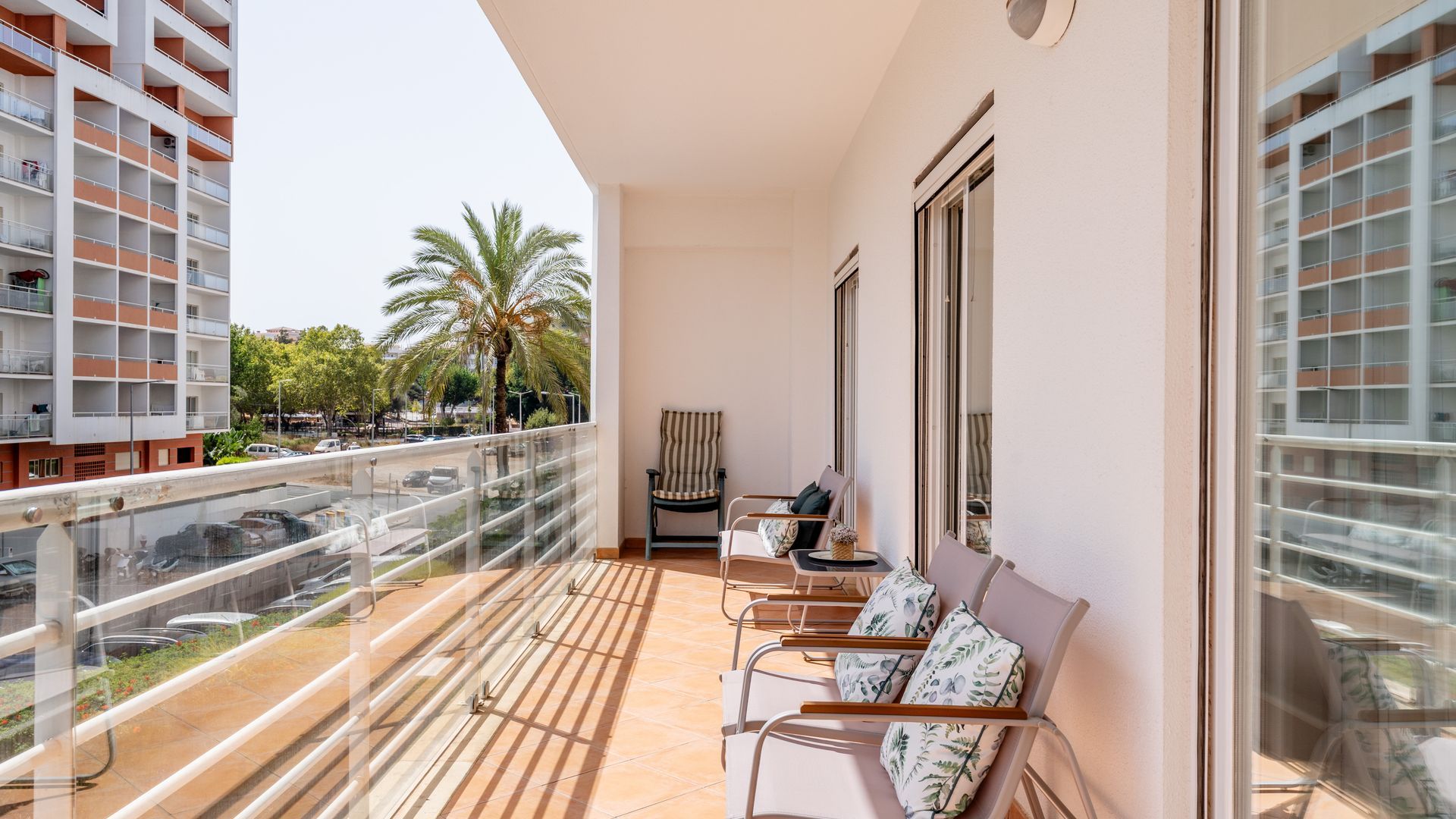 Un balcon avec vue sur un palmier et un immeuble. On y trouve deux chaises et une table avec un vase.