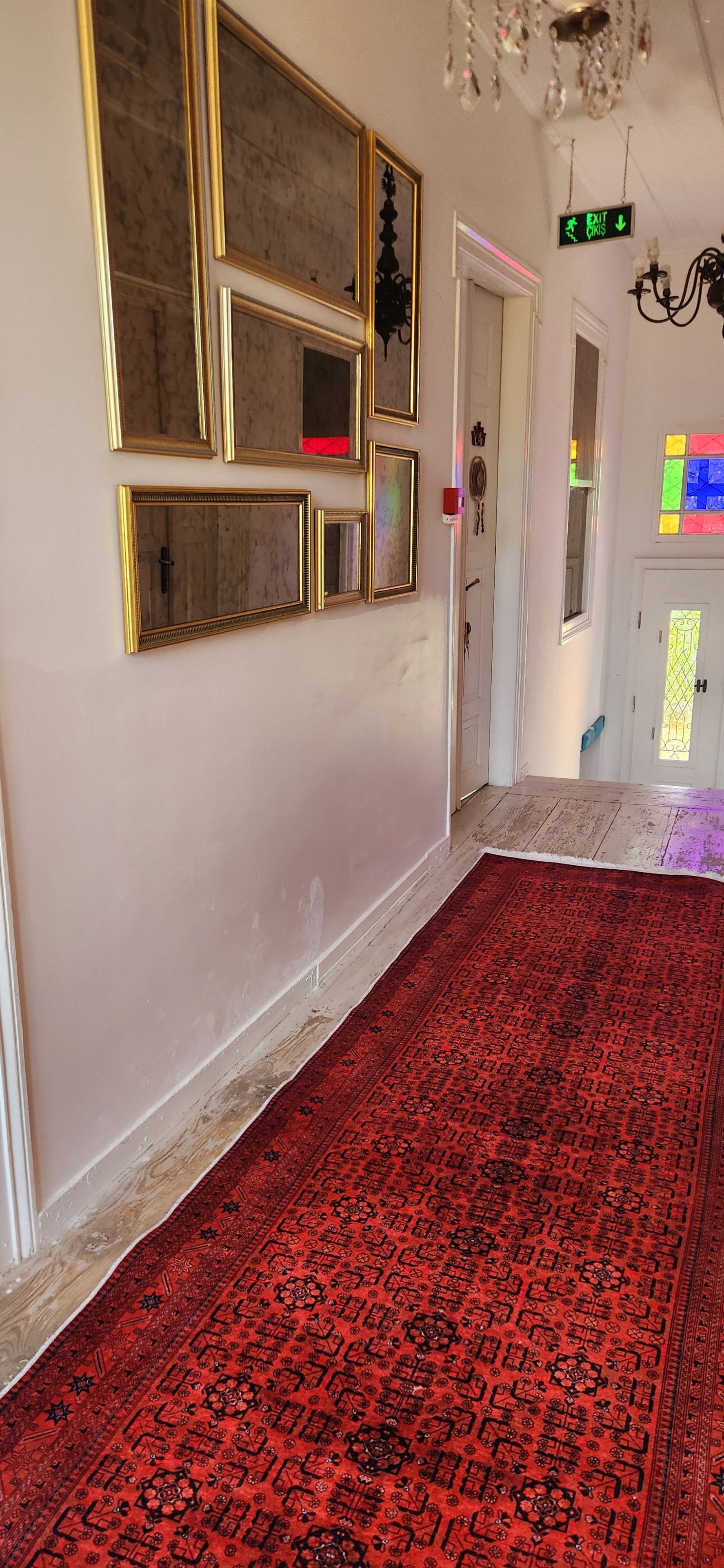 A hallway with a red patterned carpet, a wall with multiple framed mirrors, and a door with an exit sign.