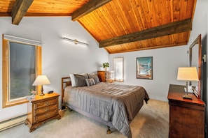 A bedroom with a wooden ceiling, a bed with a grey quilt, a nightstand with a lamp, a dresser, and a window with blinds. - Ski Ranch 204 - 2 bedroom + loft, walk to the resort! (Steamboat Springs)