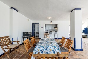 Zona de menjador amb taula llarga, cadires, una barbacoa i un espai de cuina amb armaris i un rentaplat.