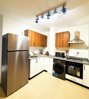 A compact kitchen with a stainless steel refrigerator, wooden cabinets, a washing machine, and a microwave. - Edelma Retreat 2BR Pool Views, Sahl Hasheesh (Red Sea Governorate)
