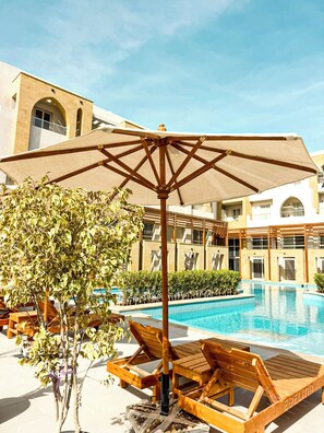 A pool area with lounge chairs, a large umbrella, and a building with arched windows in the background. - Edelma Retreat 2BR Pool Views, Sahl Hasheesh (Red Sea Governorate)