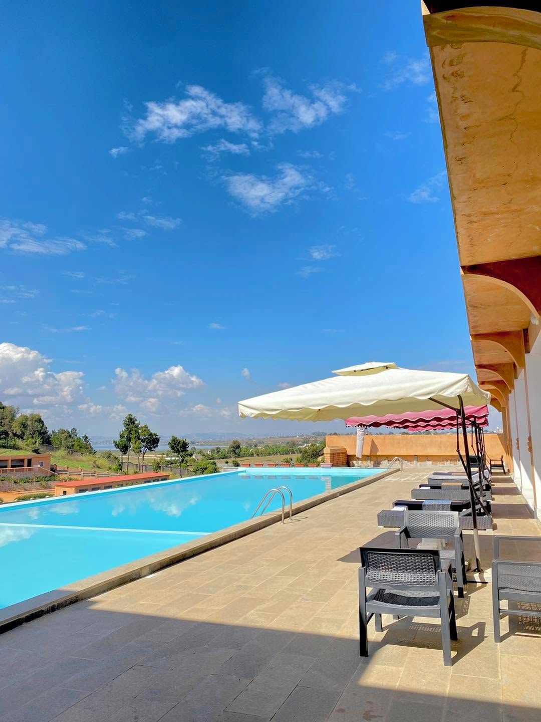 A swimming pool with lounge chairs and umbrellas, surrounded by a clear sky and distant landscape.