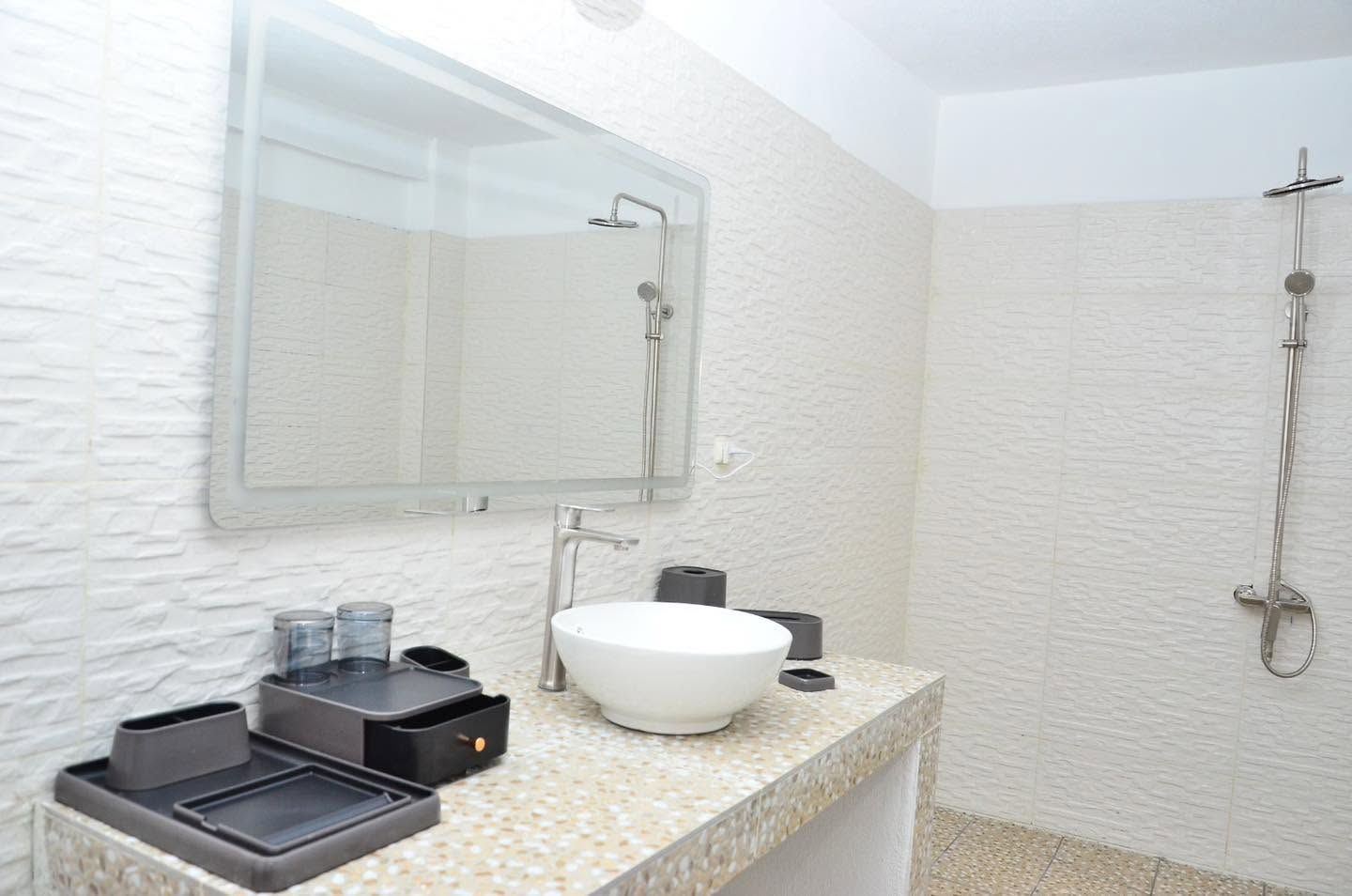 A bathroom with a white textured wall, a large mirror, a white sink, a shower, and a countertop with a trash bin and a soap dispenser.