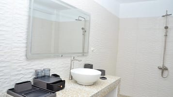 A bathroom with a white textured wall, a large mirror, a white sink, a shower, and a countertop with a trash bin and a soap dispenser.