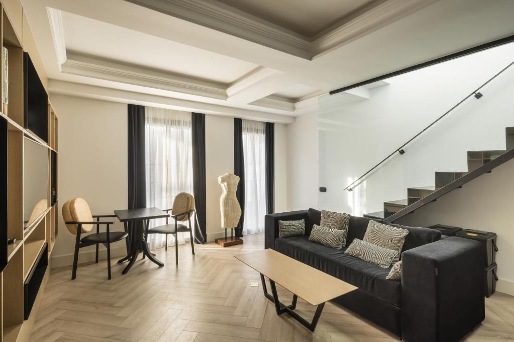 Una sala moderna con un sofá, una mesa de centro y una zona de comedor con sillas. Hay una escalera que sube a un piso superior.