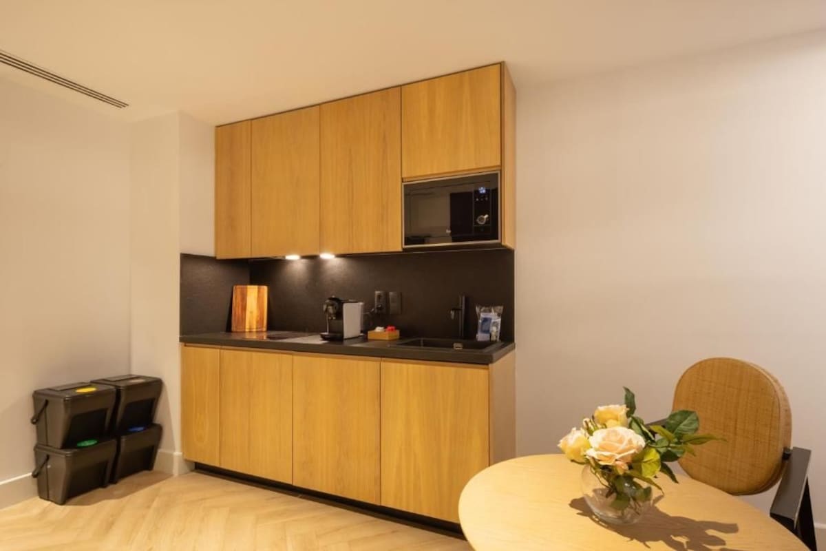 A modern kitchen with wooden cabinets, a black countertop, and a microwave. There is a small table with a vase of flowers.
