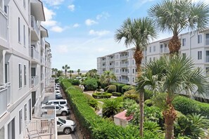 A multi-story residential building with balconies, a parking area, and well-maintained greenery including palm trees and shrubs.