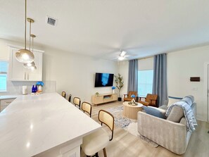 A modern living room with a sofa, coffee table, and TV. There is a kitchen area with a dining table and chairs. - Azure & Anchor Coastal Escape W Huge Fenced Yard & Fire Pit + Small-Pet-Friendly (St. Augustine)