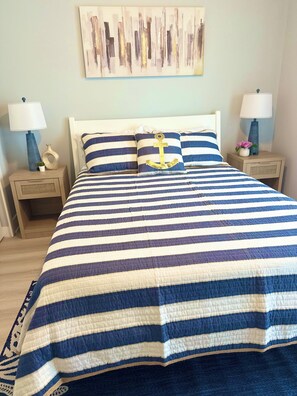 A bed with a striped bedspread, two bedside tables with lamps, and a wall art piece above the bed. - Azure & Anchor Coastal Escape W Huge Fenced Yard & Fire Pit + Small-Pet-Friendly (St. Augustine)