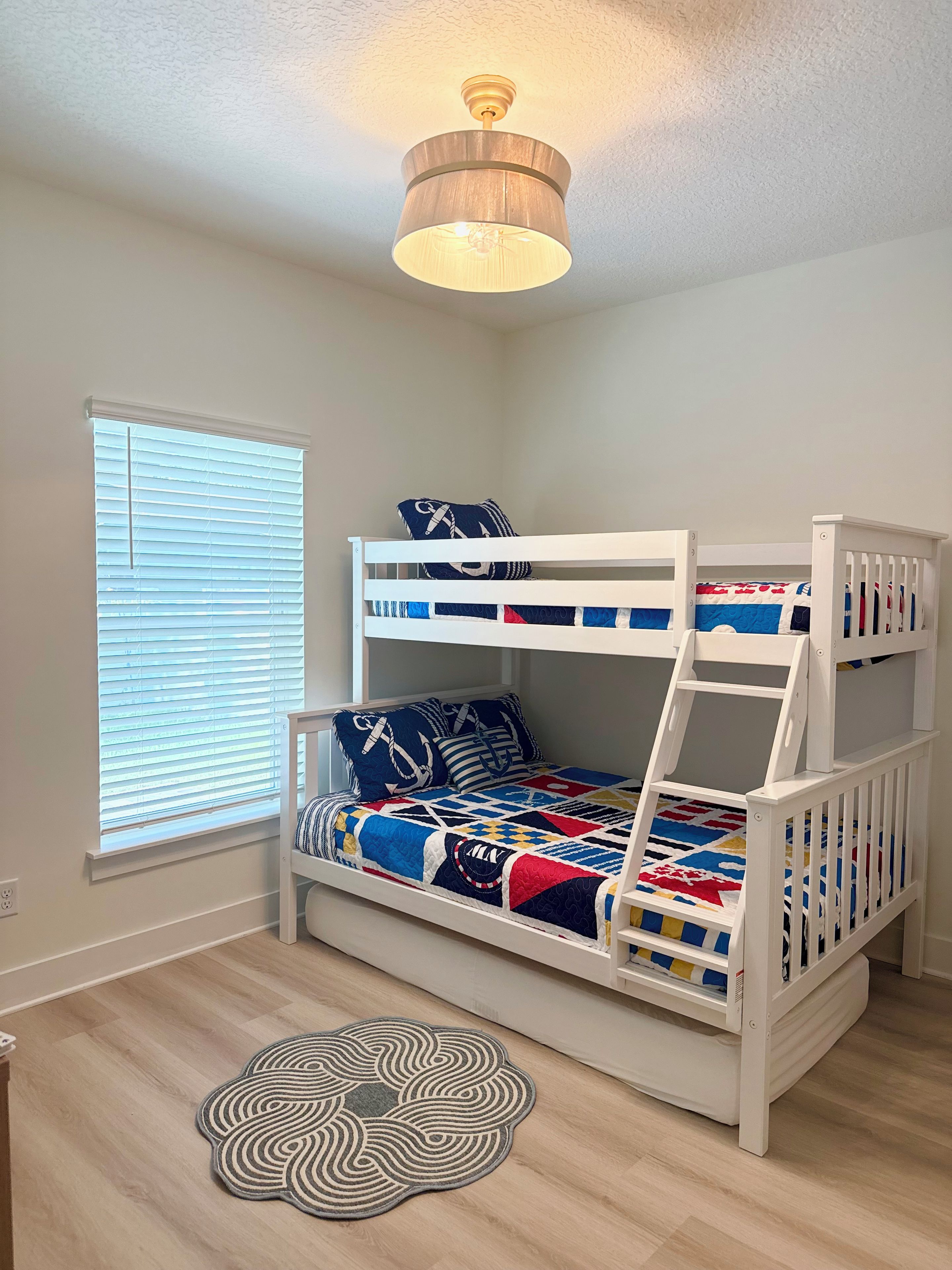 A bunk bed with a nautical-themed bedding, a ceiling fan, a window with blinds, and a decorative rug.