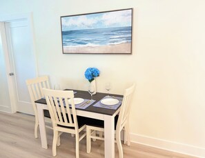 A dining area with a table set for two, a vase with blue flowers, and a framed picture of the ocean on the wall. - Azure & Anchor Coastal Escape W Huge Fenced Yard & Fire Pit + Small-Pet-Friendly (St. Augustine)