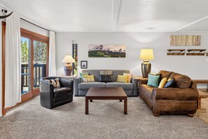 Sala de estar com sofá, poltrona, mesa de centro e luminárias. Há um quadro na parede e uma janela com cortinas.