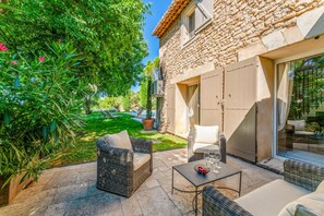 A patio with wicker furniture, a table with glasses, and a stone building with a garden. - Villa Les Vignes - Four Bedroom Villa, Sleeps 10 (Joucas)