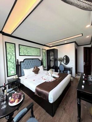 A hotel room with a large bed, a bedside table with a bottle and glasses, a small table with fruit and a wine glass, and a mirror on the wall. - vincentluxury (yên nghĩa)