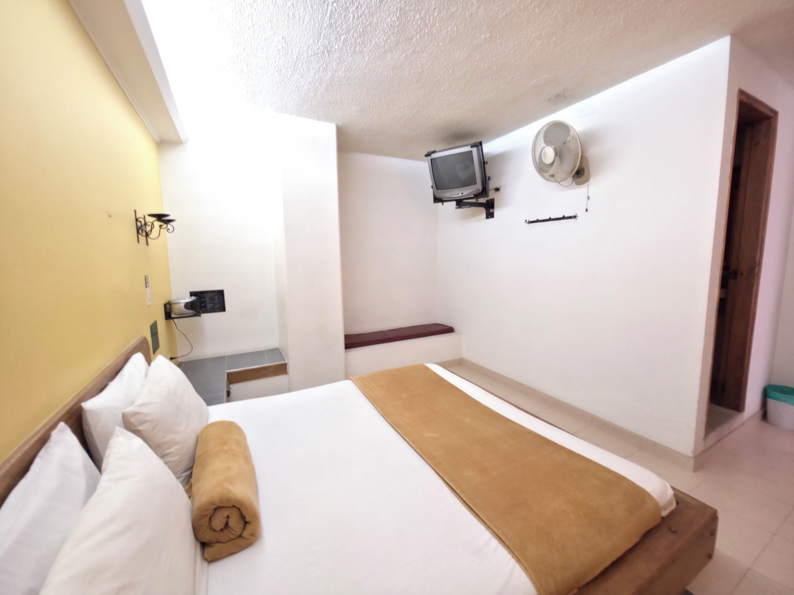 A hotel room with a bed, a television mounted on the wall, a fan, and a small seating area.