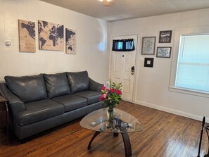 Ein Zimmer mit einer schwarzen Ledersofa, einem Glastisch mit Blumen in einer Vase, Holzböden, einer weißen Tür und Wanddekorationen, darunter Weltkarten und motivierende Zitate.